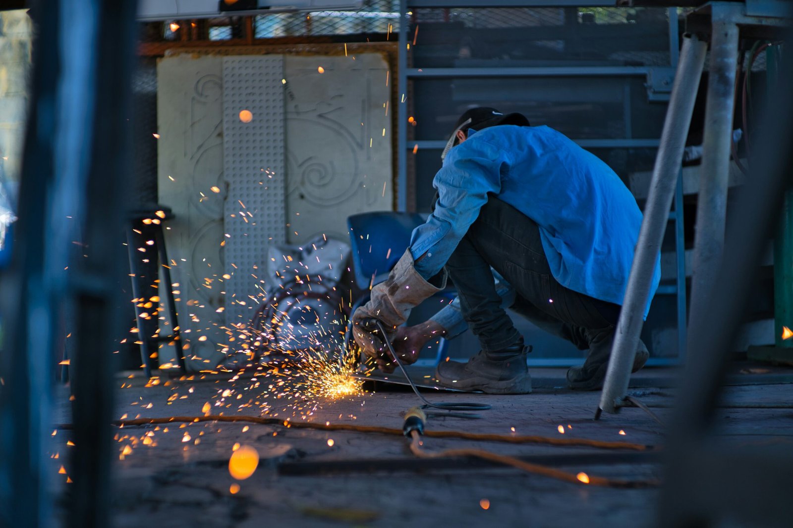 A worker in a blue shirt and protective gloves crouching on the ground while welding metal, surrounded by flying sparks in an industrial setting.