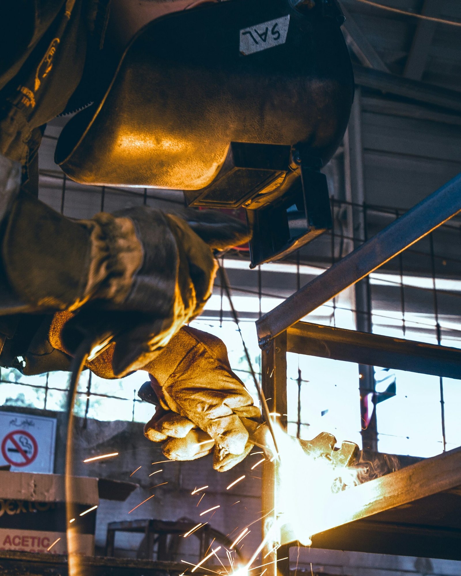 Welder in protective gear working on a metal frame indoors, with flying sparks and welding mask visible.